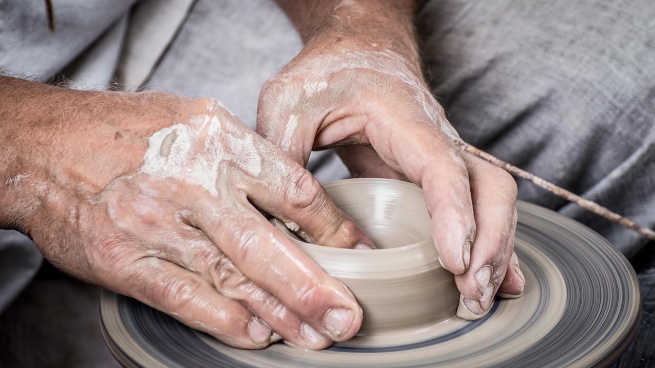 Clay-covered hands shape a small pale vessel on a spinning pottery wheel.