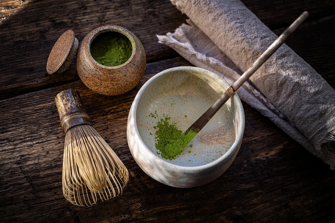 A pale ceramic tea bowl with green powder sits beside a whisk, scoop, cloth, and textured containers on dark wood.